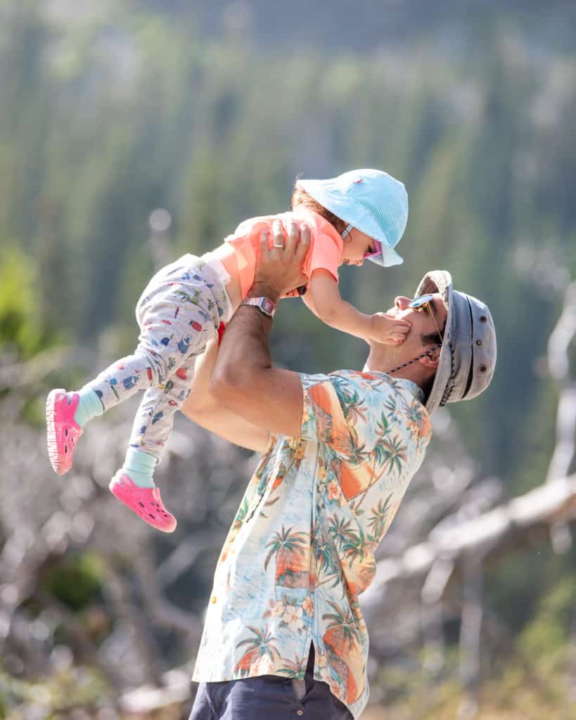 father holding toddler daughter up in the air. Both are wearing hats and sunglasses.