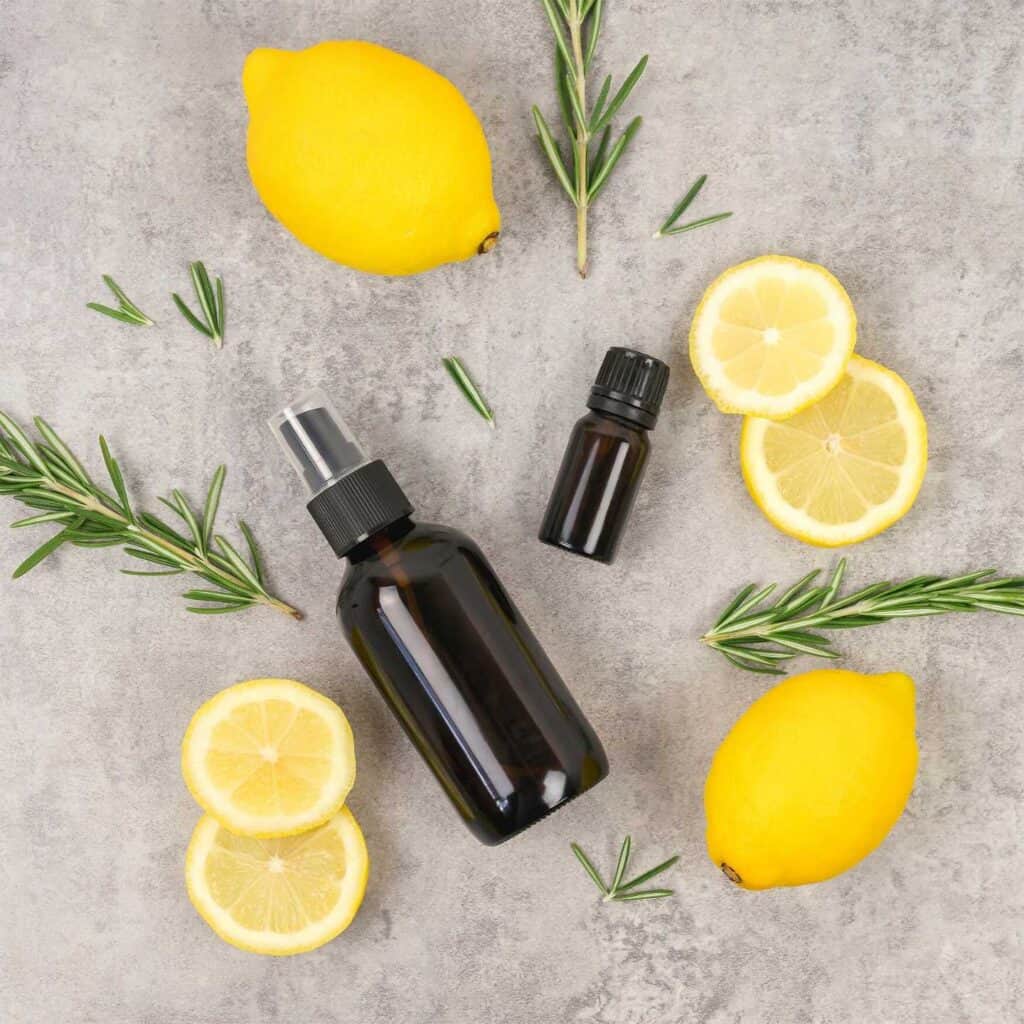 an amber glass 2 ounce spray bottle and a small essential oil bottle lie on a table surrounded by lemon and rosemary