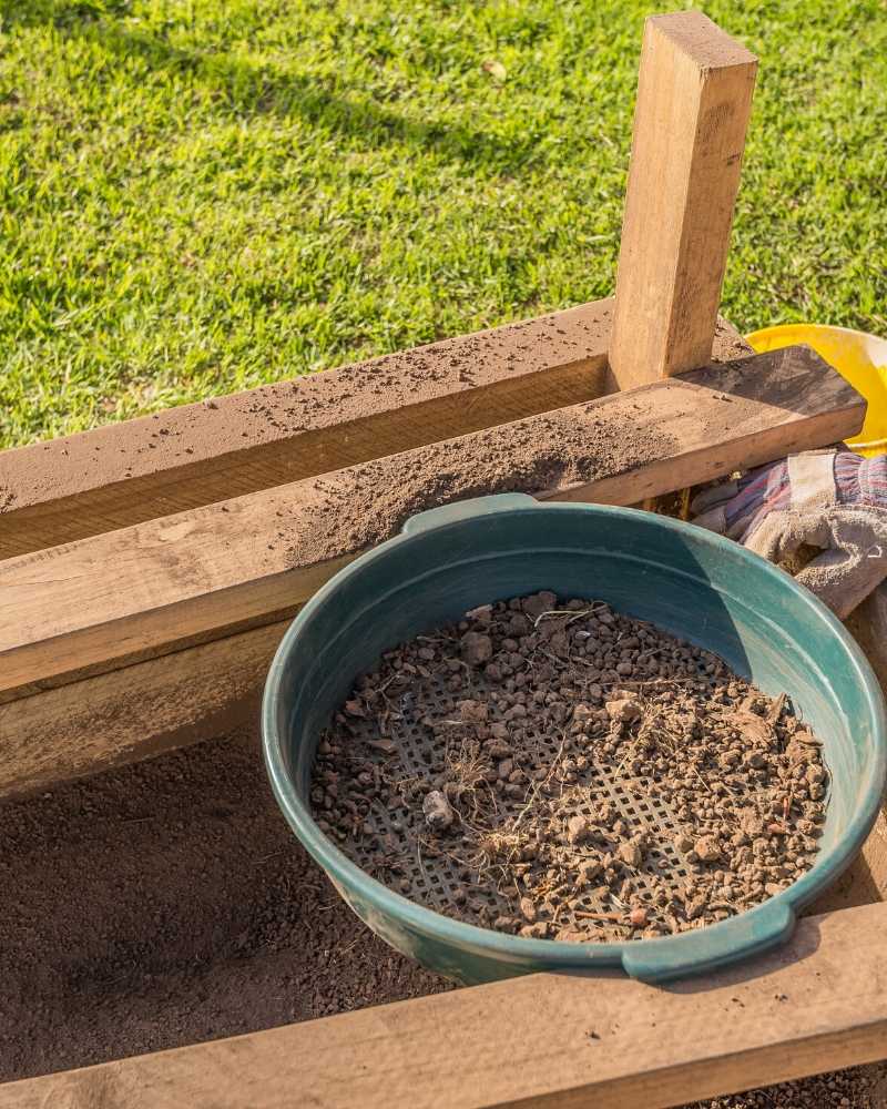 a large green garden sieve with a small amount of dirt in it. the garden sieve is resting on a 2x4 frame and underneath it is a wooden container with sifted soil