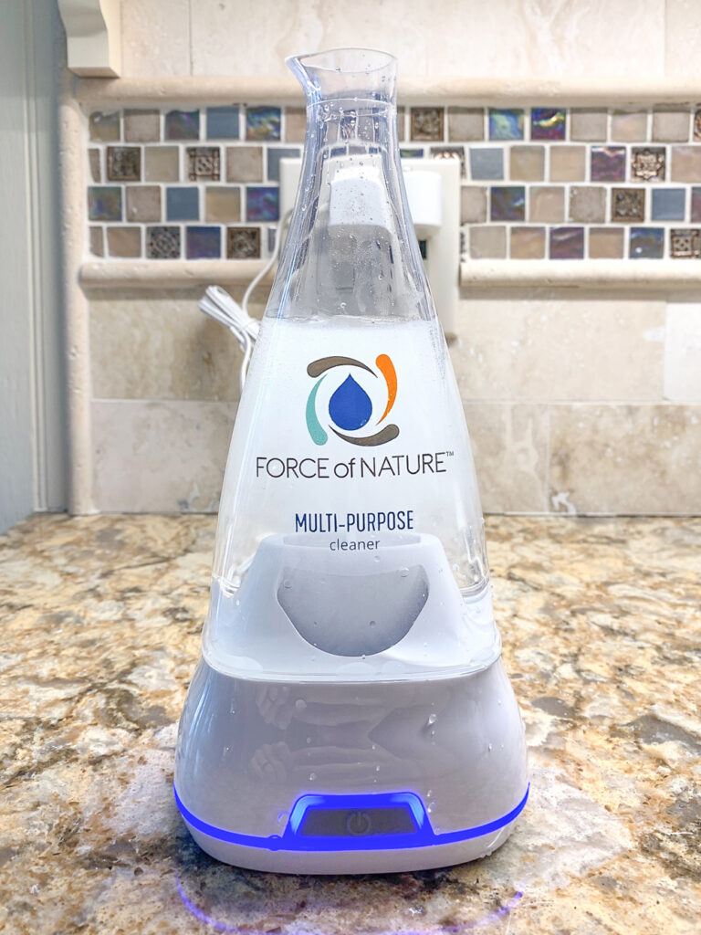 An electrolyzer of Force of Nature multipurpose cleaner on a tan silestone counter. A tan tile backsplash is visible in the background. The electrolyzer's base has a blue light and the liquid visible inside is white from small bubbles and the electrolyzing process.  