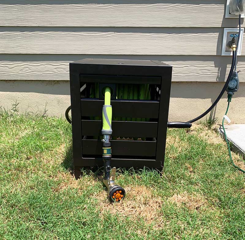A metal hose reel cabinet on the ground in front of a tan house wall. The hose reel cabinet has a birhgt green hose.