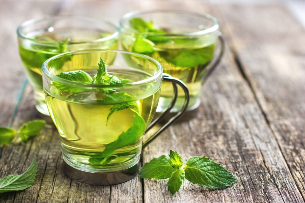 three glass teacups with fresh mint tea on a wood table. There are fresh mint leaves in each cup and on the table.