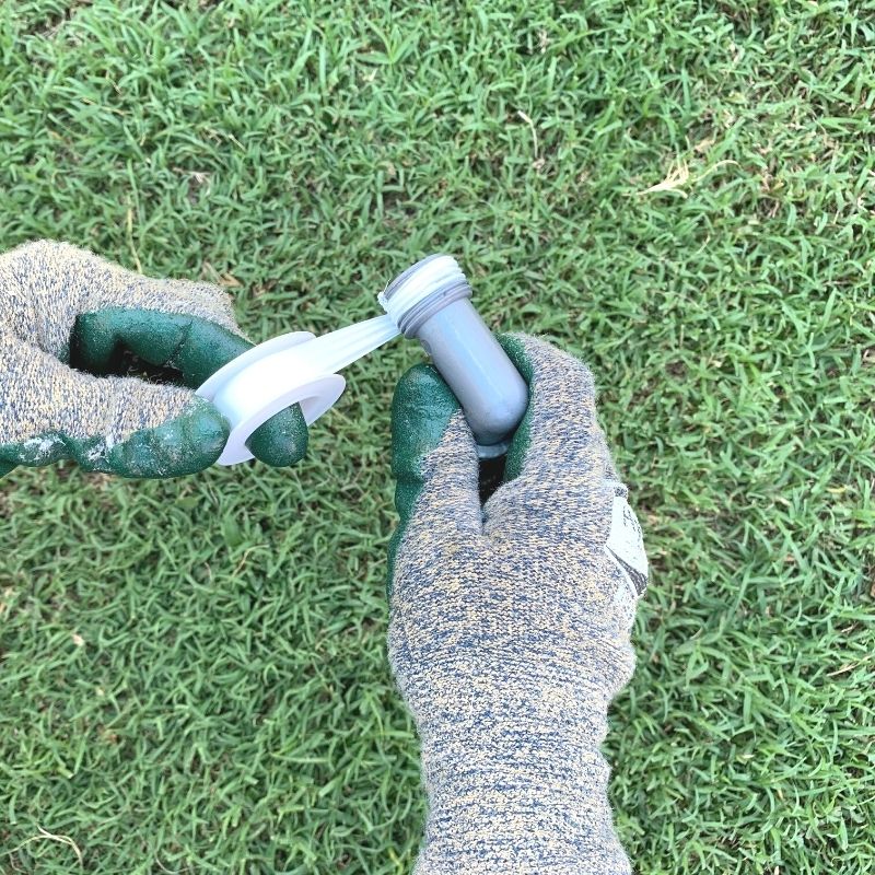 Gloved hands wrapping a male hose reel cart union with plumber's tape. The gloves are green and there is grass in the background 