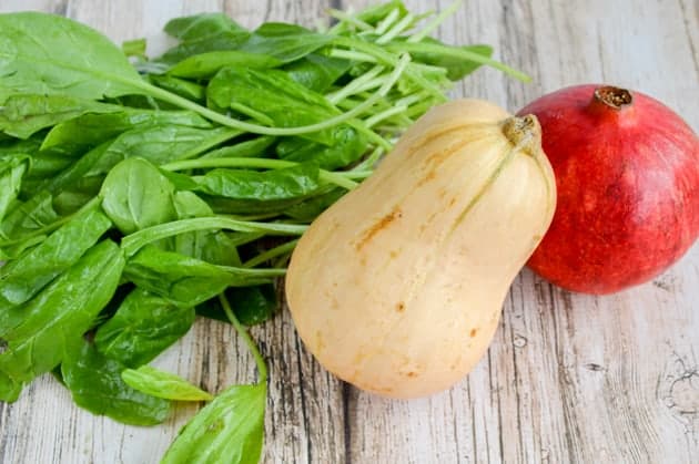 a bunch of spinach leaves, a whole butternut squash, and a pomegranate on a light wood background