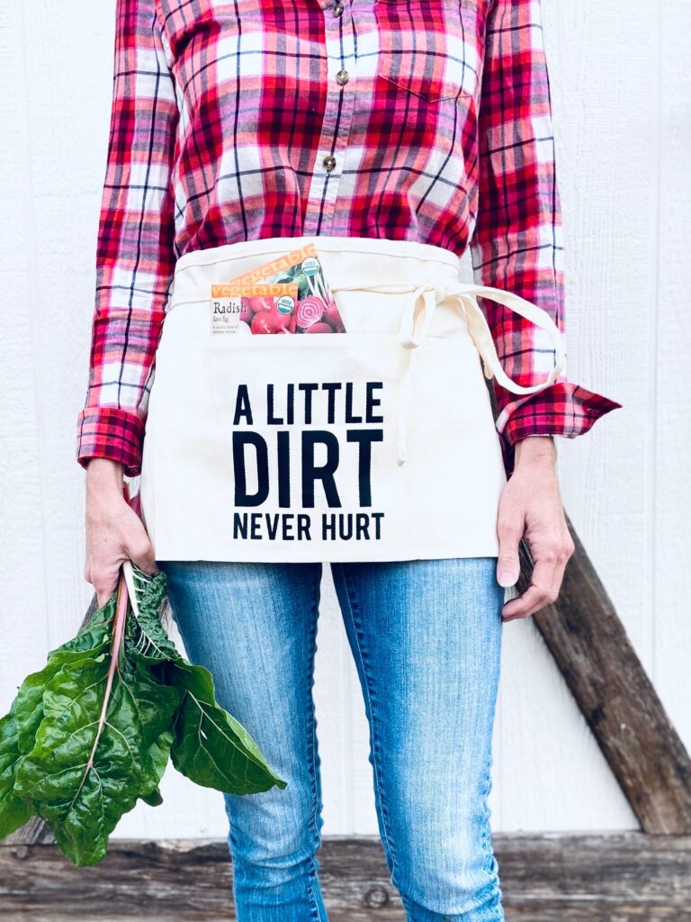 A woman holding swiss chard and wearing a waist apron with the text "a little dirt never hurt." There are radish seed packets sticking out of the apron. She is wearing a red flannel shirt and jeans. The photo shows from her knees to her collar bone - her feet and face are not in the frame.