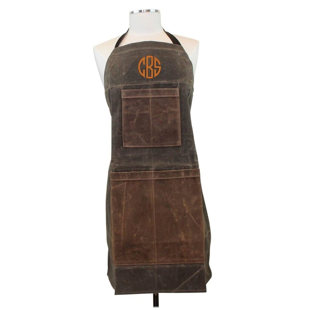 A long brown waxed canvas apron with an orange monogram "CBS" on the chest. The apron is on a plain dress form without a head or arms.