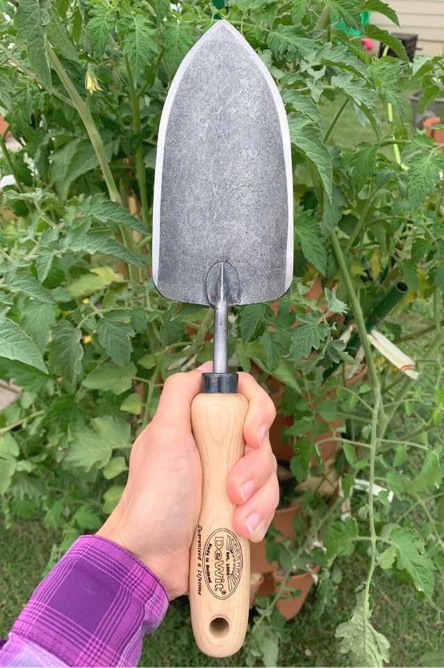 a photo of a woman's hand holding a DeWit brand gardening trowel against a backdrop of tomato plants. 