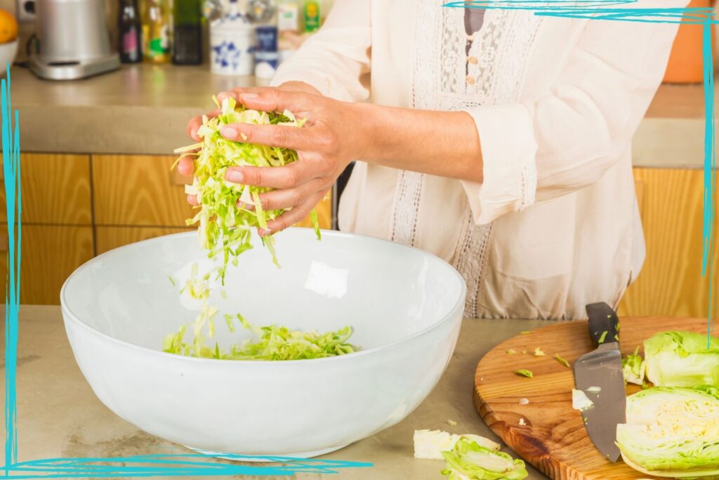 A close up shot of a woman in a kitchen shredding cabbage for coleslaw or sauerkraut. She has a large white bowl full of shredded cabbage and a handfull of cabbage she's dropping into the bowl