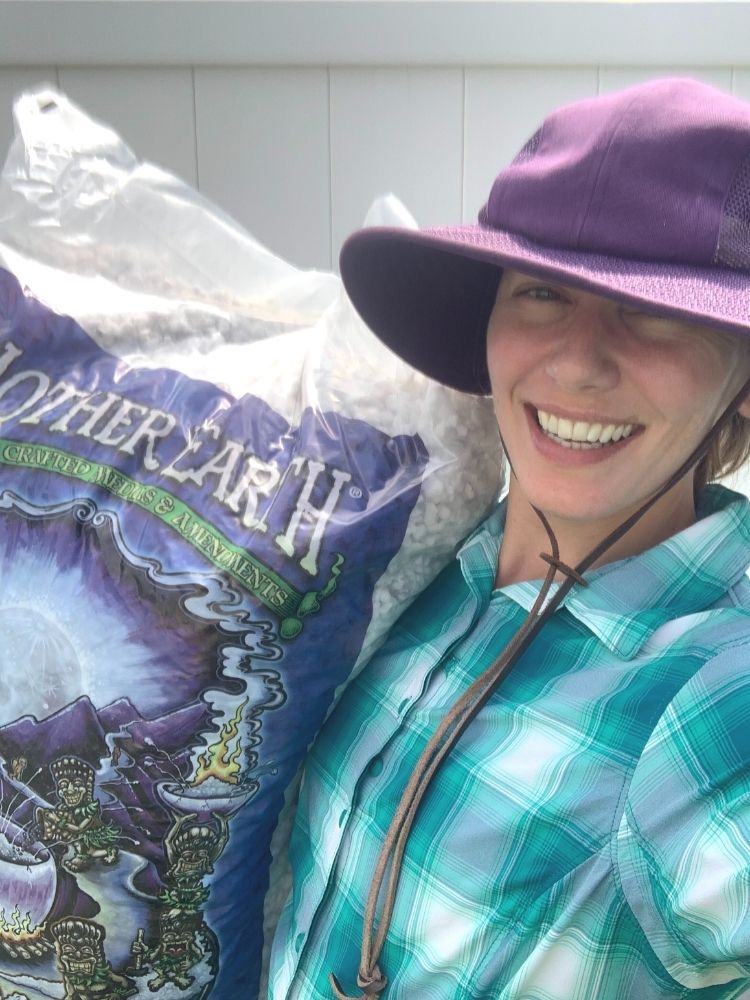 A selfie of a woman wearing a teal plaid shirt and a purple gardening hat holding a large bag of perlite