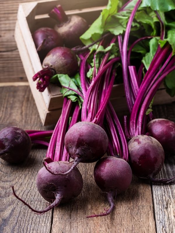 A bunch of harvested beets in a wood crate. The roots are round and red and the beet leaves are still attached