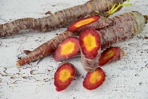 An image of two whole purple carrots and one sliced purple carrot with a yellow center. The carrots on are a weathered white wood surface.