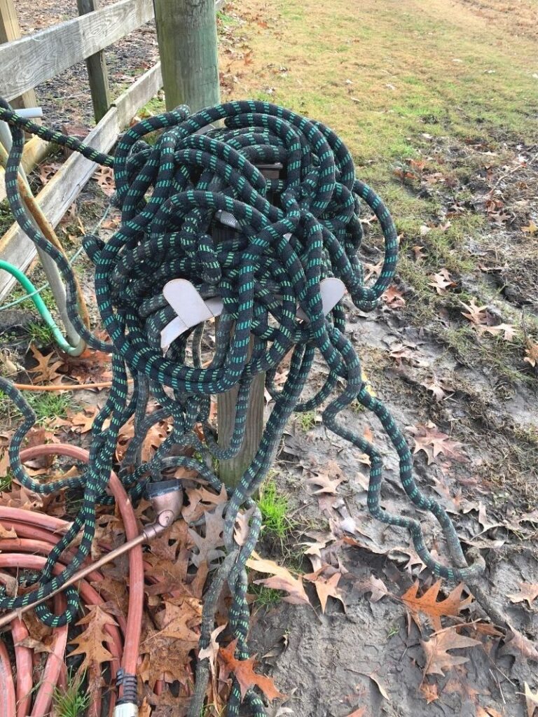 A long, tangled expandable hose heaped on top of a post and falling onto muddy ground. Also visible on the ground is a red garden hose with a brass colored watering wand. 