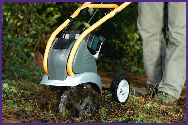 A close up of a person wearing long pants and boots using a cultivator on a patch of ground 