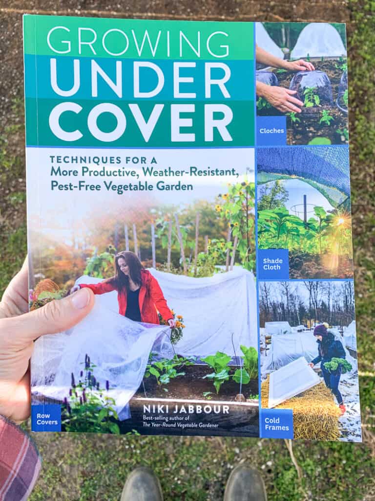 A hand holding the gardening book "Growing Under Cover" by Niki Jabbour