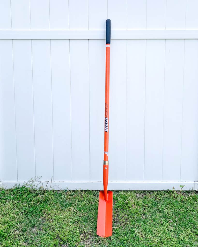 An orange trenching shovel with an orange fiberglass blade leaning against a white vinyl fence