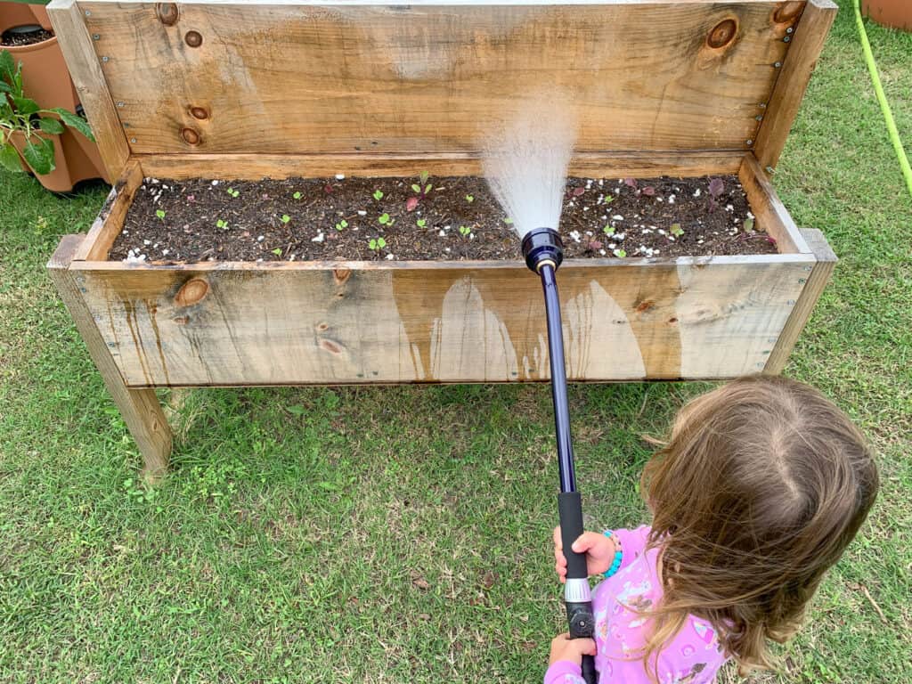 A young child in bright pink pajamas using a watering wand to ater a raised planter.