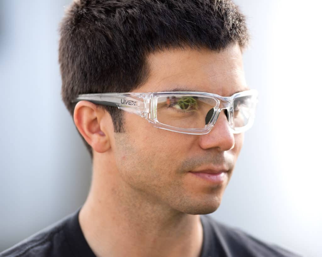 Photo of a man wearing the clear UVEX safety glasses looking away from the camera. Background is an out of focus white fence.