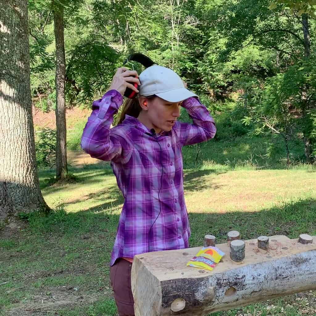 Photo is of a woman placing a set of earmuffs over a set of earplugs and showing how to wear double hearing protection. The woman is a wearing a purple plaid shirt, red pants, and a white hat. The background is of a wooded lot.