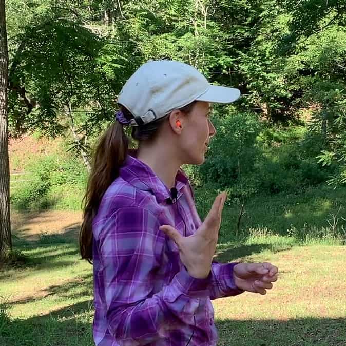 Photo is of a side profile of a woman wearing orange earplugs. She is wearing a purple plaid shirt and a white hat. The background is a forested lot.