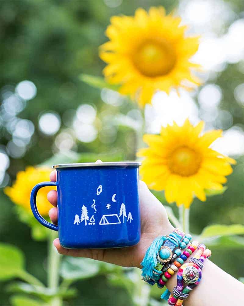 A hand holding a blue camp mug in front of a garden patch with blooming sunflowers