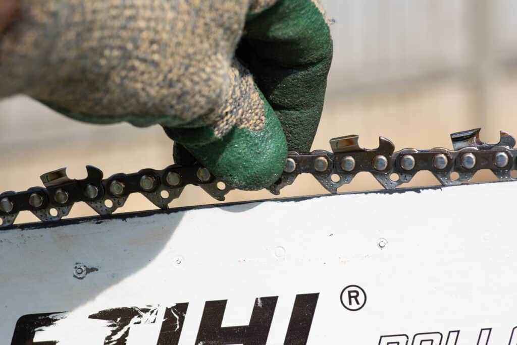 Photo is of a loose chain on a chainsaw. A gloved hand is pulling up on the chain of a chainsaw and the drive links are exposed as the chain pulls away from the guide bar.