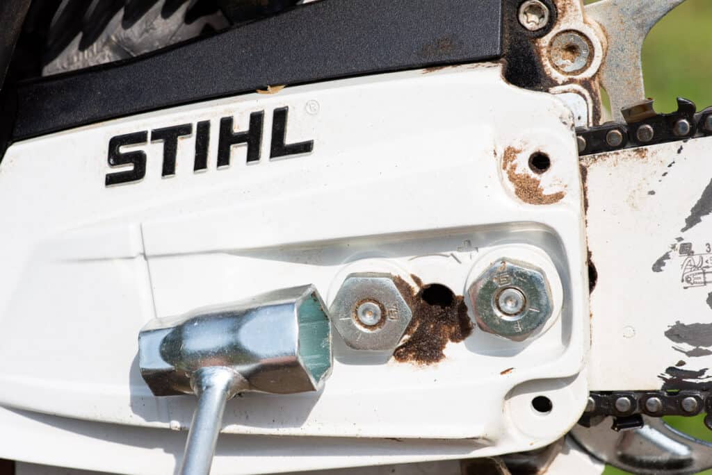 Photo is of a side profile of a Stihl chainsaw. A chainsaw multi tool is shown in front of the side panel nuts to show which nuts need to be loosened to tighten the chain.
