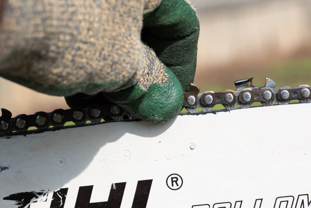 Photo is of a tight chain on a chainsaw. A gloved hand is pulling up on a chainsaw chain. The drive links are barely exposed showing that the chain is tight.