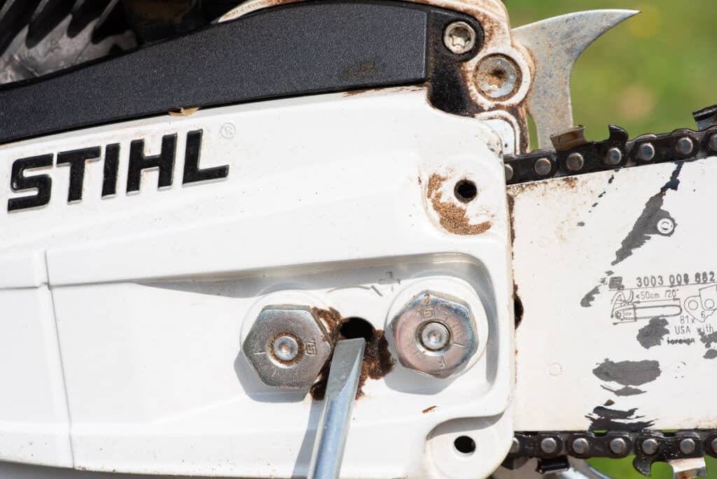 Photo is of a side profile of a chainsaw. A flat head screw driver is being inserted into the tightening port to tighten the chain on a chainsaw.