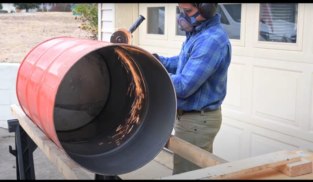 Photo is of a man cutting a 55 gallon drum in half using an angle grinder. The barrel is on 2 2x4s so it does not rock back and forth.