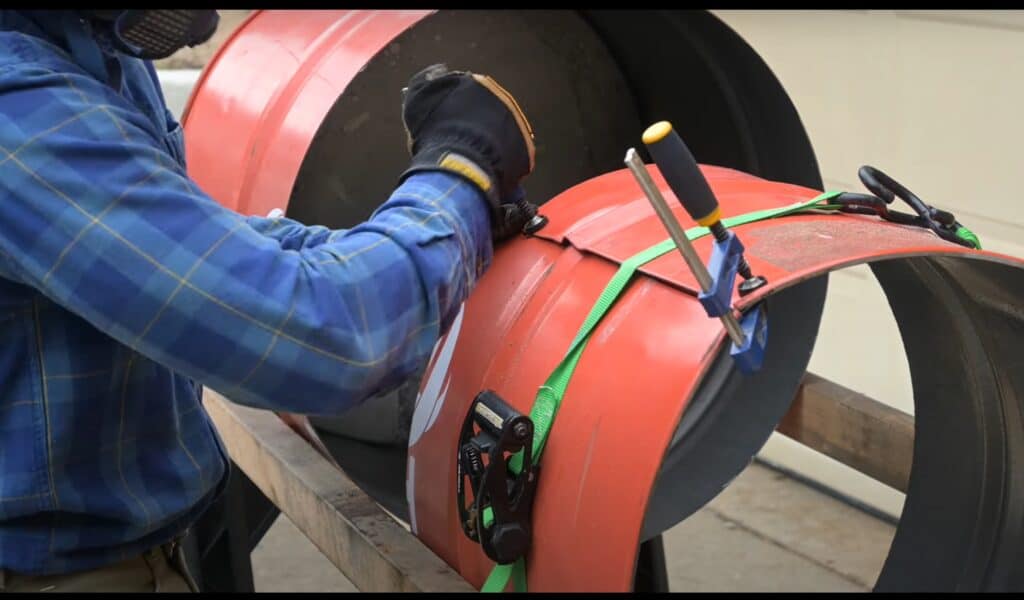 Photo is a of a man folding a 55 gallon drum to reduce the diameter of the barrel.