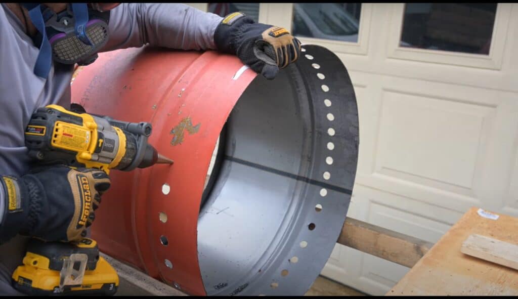 Photo is of a man drilling 3/4" holes from the outer shell of a 55 gallon drum firepit.