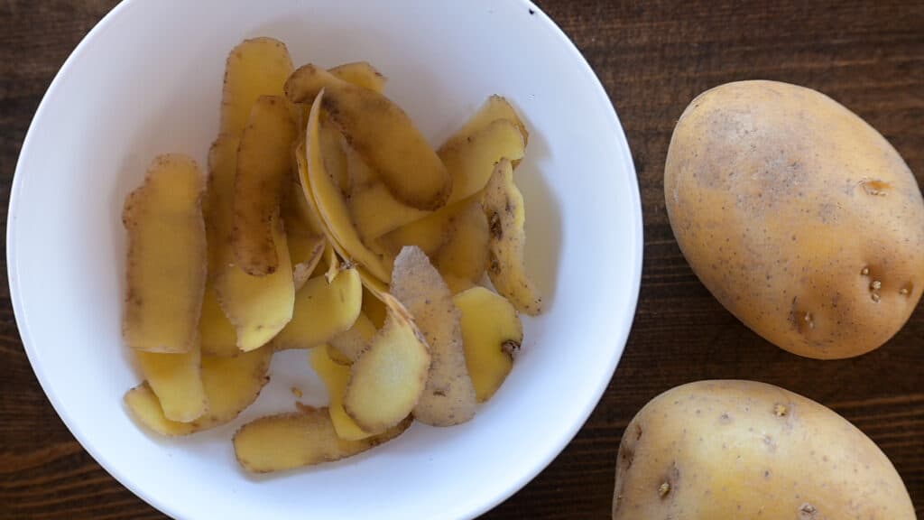 A picture with a bowl of potato peels next to two yellow ptoatoes