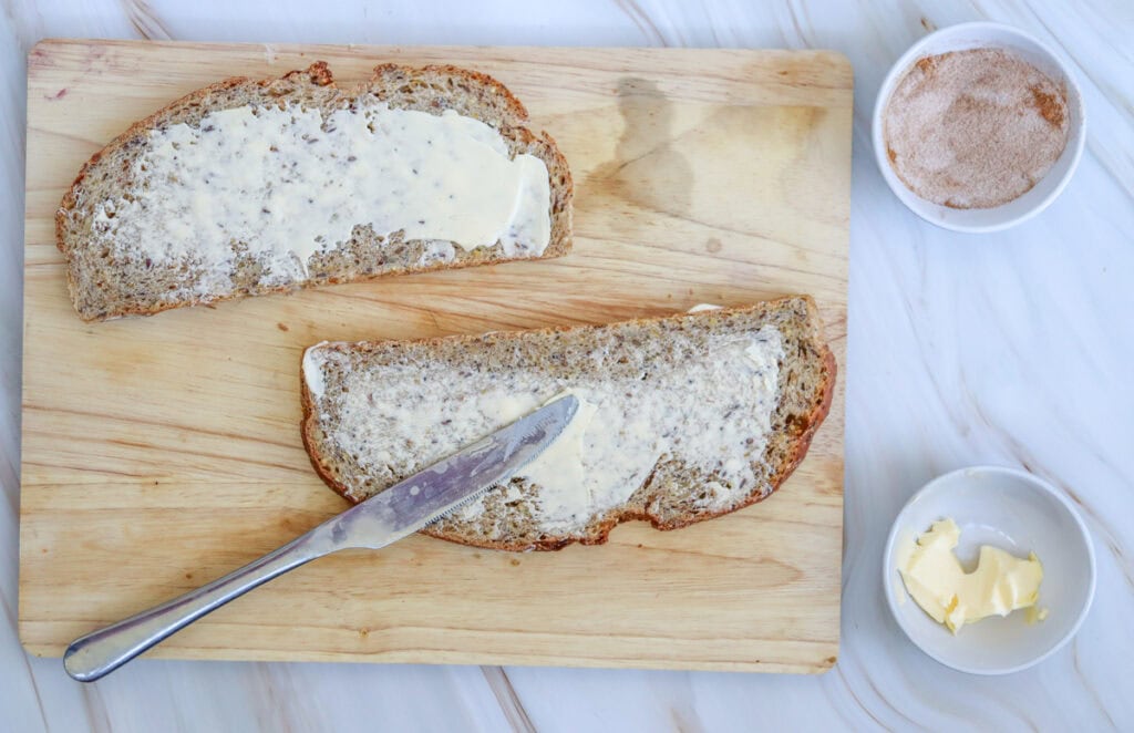 A wooden cutting board displays two slices of sourdough bread spread with butter, accompanied by small bowls of cinnamon sugar and more butter, suggesting ideas for breakfast sourdough toast toppings.