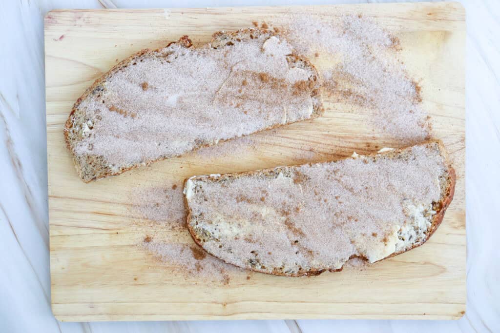 Two slices of sourdough toast, topped with a mixture of cinnamon and sugar, are placed on a wooden cutting board.