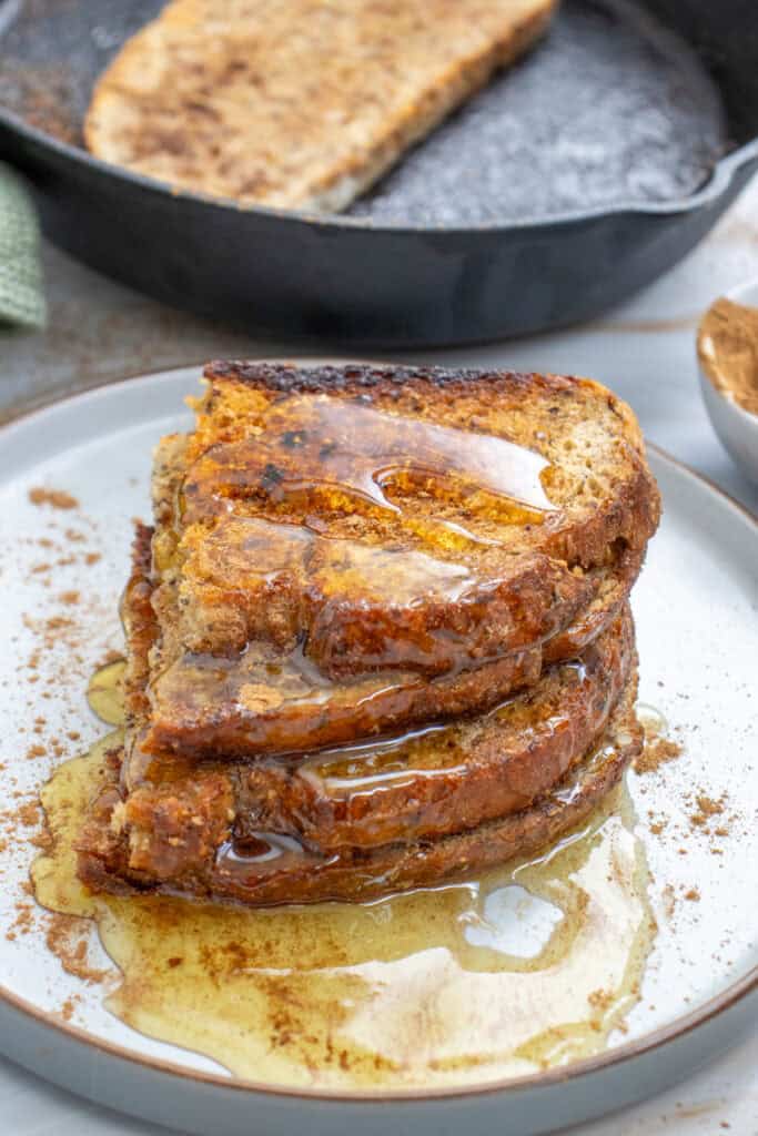 A stack of sourdough toast drizzled with honey sits on a plate with a small bowl of cinnamon in the background, suggesting sweet sourdough toast ideas for breakfast.
