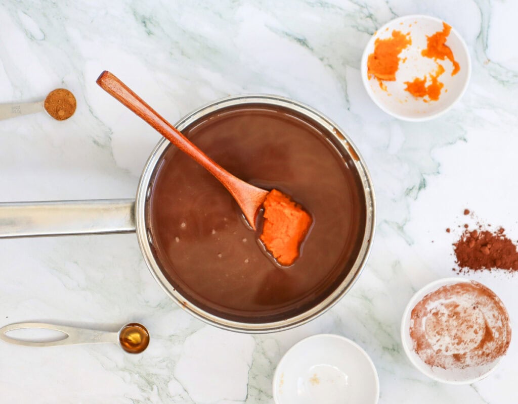 A stainless steel pot filled with chocolate liquid is stirred with a spatula, with bowls containing pumpkin puree and cocoa powder arranged nearby, suggesting a preparation for fall pumpkin hot chocolate recipes.
