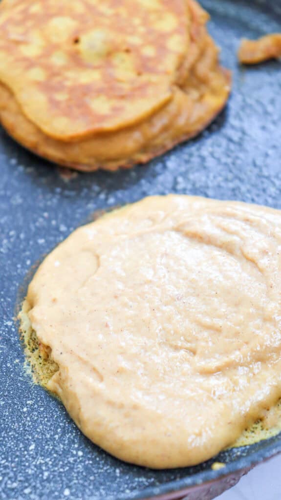 A skillet contains a scoop of pumpkin pancake batter cooking alongside a finished pumpkin pancake, illustrating a quick pumpkin pancake recipe for easy autumn breakfasts.

