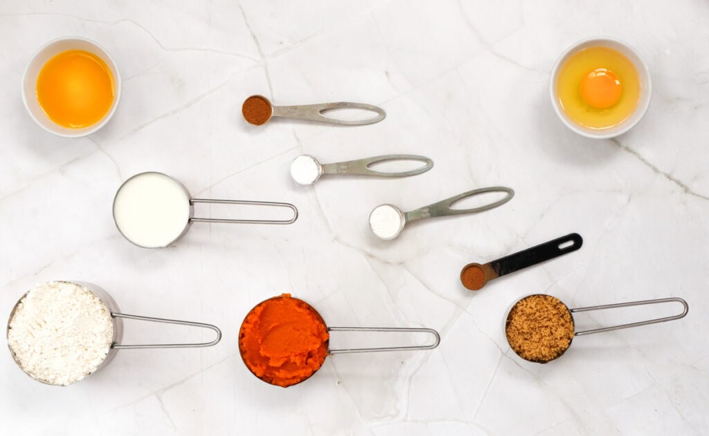 An overhead view of ingredients arranged on a marble surface, including eggs, milk, flour, pumpkin puree, brown sugar, and various spices, for a quick pumpkin pancake recipe.

