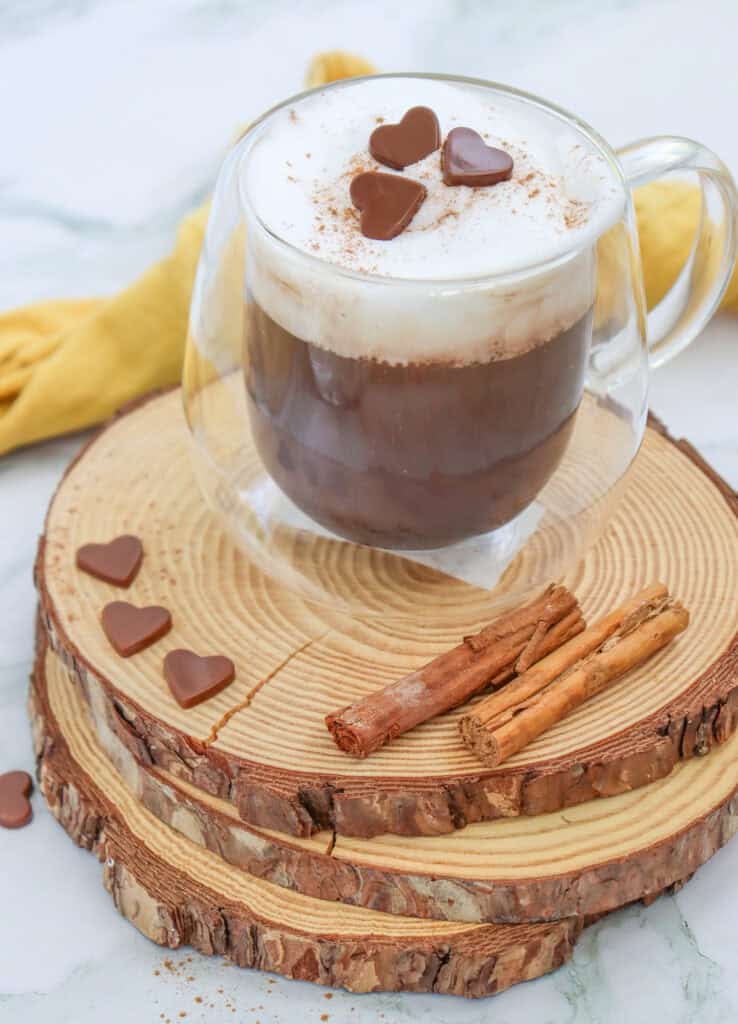 A clear glass mug filled with hot chocolate topped with whipped cream, chocolate hearts, and a sprinkle of cinnamon sits on a wooden platter alongside cinnamon sticks, surrounded by a yellow cloth and suggesting fall hot chocolate recipes.

